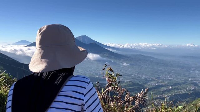 Slow motion shot from behind of a woman wearing a hat enjoying the breathtaking view of Mount Sindoro, Sumbing, and a sea of clouds from the summit of Mount Bismo in Indonesia.