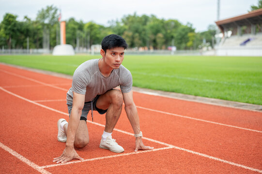 Asian male sprinter crouching in low start position on running track line in sports stadium.