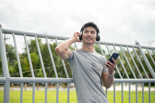 Asian athletic man listening to music with wireless headphones while using smartphone outdoors