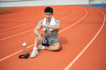 Obraz premium Asian athletic man checking time or reviewing smartwatch while recovering on a track after running.