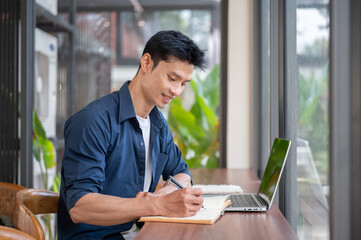 Fototapeta premium Asian man writing notes while working or study on laptop at wooden counter table in cafe