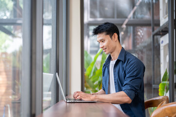 Fototapeta premium Young Asian freelancer working on laptop at wooden table beside glass wall in modern cafe