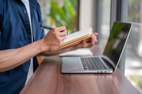 Close up of a freelancer or man student writing notes beside laptop while working at table in cafe.