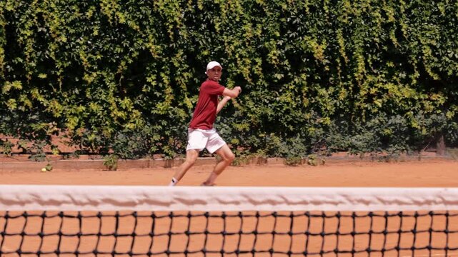 Male tennis player hitting two handed backhand across net on outdoor clay court. Concept of competitive mindset, tournament season, player development, coaching programs and sports education.