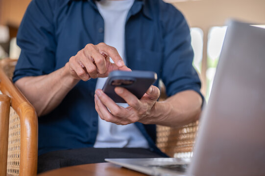 Close up of a freelancer adult man's hand using smartphone beside laptop while sitting at cafe table