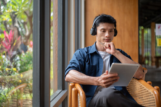 Focused Asian businessman wearing headphones using digital tablet while working at table in cafe.