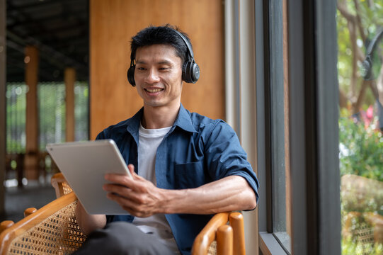Asian freelancer man smiling while using a tablet and wearing headphones working in the cafe.