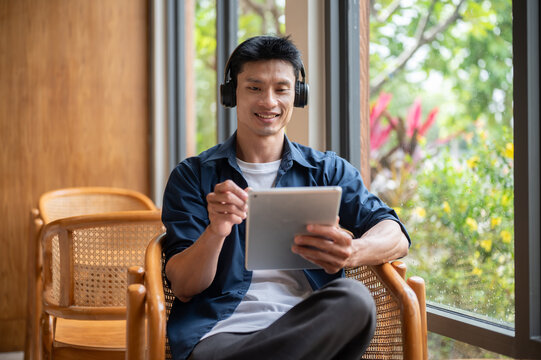Asian artist man using a tablet and enjoy listening to music in headphones while working in the cafe