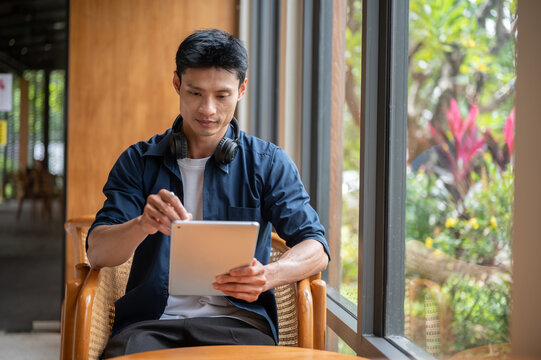 Asian artist man using a stylus to write on a tablet while sitting at cafe table beside glass wall.