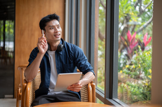 Asian freelancer man holding a tablet with thinking gesture while sitting in modern cafe.
