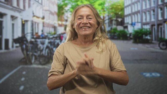 Senior woman clapping hands and raising arms on a cobbled city street lined with bicycles and row buildings; joy celebration.