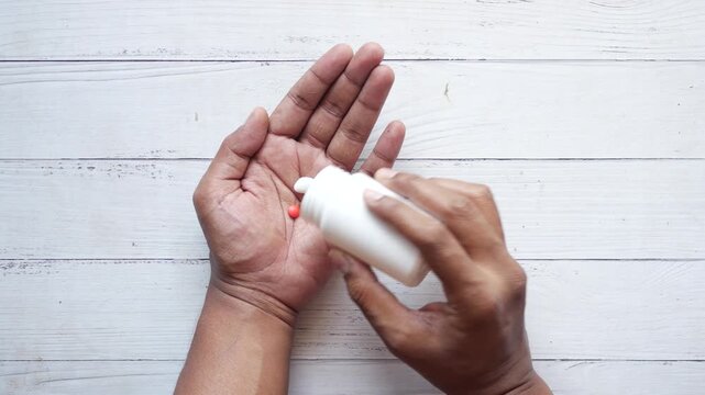 A person pouring different colored pills from a white bottle onto their outstretched palm, captured from a top-down perspective showing the action and detail of the medication