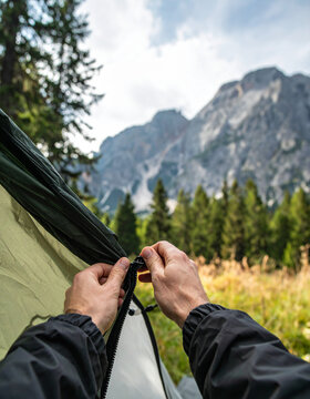 Close-up of hands zipping up a tent at a scenic campsite surrounded by tall pine trees and mountain peaks.