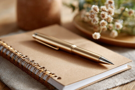 Matte gold ballpoint pen resting on kraft paper spiral notebook with dried flowers in warm workspace