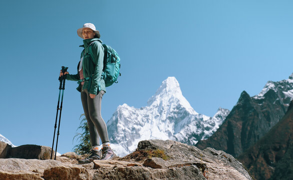 Active woman hiker with backpack and trekking poles standing on rocky trail with snowy mountain peak on background with Ama Dablam in Nepal on background.