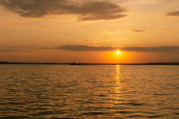 Obraz premium Golden sunset over calm water with reflections and gentle waves. Orange sun setting down above peaceful sea horizon with low clouds, sunny path and small distant island for nature landscape concept.