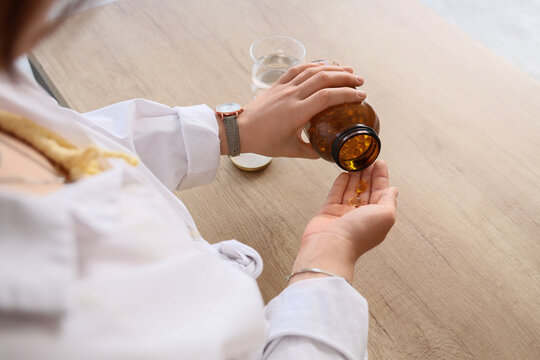 Woman pouring vitamin supplements from a brown bottle into her hand on a wooden table with a glass of water, a glass of water is visible next to the bottle