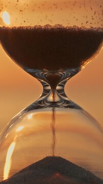 Hourglass sand trickling down close up vertical 9:16 mobile phone format warm sunset background golden hour lighting time management passing concept.