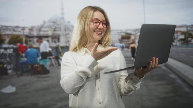Woman wearing red glasses holding laptop with palm up presenting and smiling on street by mosque and bridge; confidence connection.