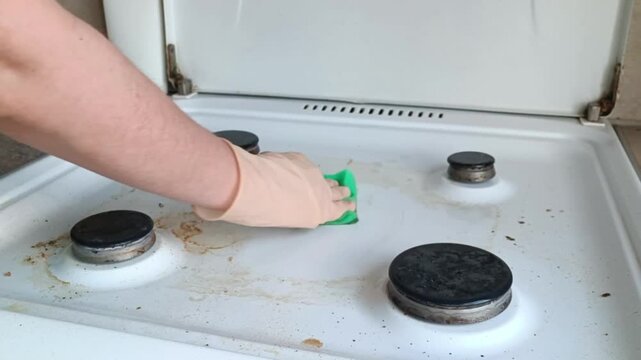 Gloved hand scrubbing a heavily soiled gas stove with a green sponge, removing grime and food splatters during deep kitchen cleaning.