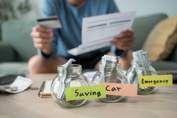 Fototapeta na wymiar Man checking financial bills and holding credit card with money jars labeled for saving, car, and emergency fund on the table, showing smart personal budgeting