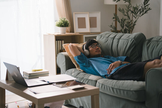 Man sleeping on a couch with headphones on after working on a laptop on the table, showing remote job exhaustion, afternoon nap, and home relaxation