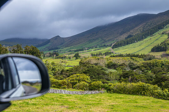 Perspectiva de un viaje por carretera con el espejo retrovisor del coche enmarcando el camino y las verdes colinas, prados y monta&ntilde;as nubladas de la isla de Azores.