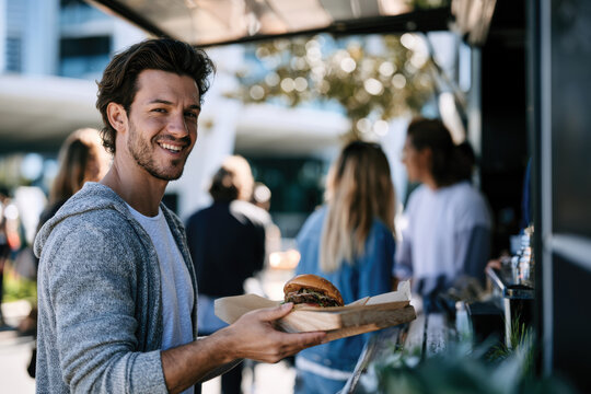 A cheerful young man stands holding a delicious burger in front of a food truck, enjoying the vibrant atmosphere filled with people in a sunny outdoor setting.