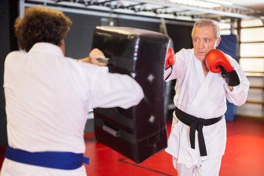 Elderly man in white kimono kicks and punches a punching bag held by a trainer