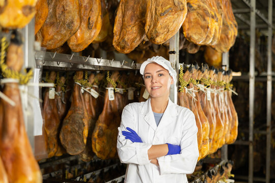 Girl employee in white uniform, bathrobe and hat, stands in workshop for production of ham. Working moments, close-knit team, private enterprise