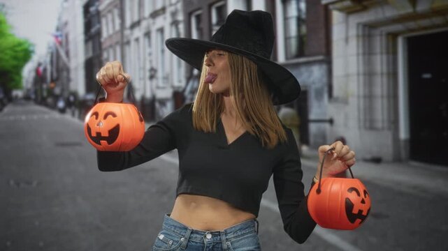 Woman wearing witch hat holding two jack o lantern buckets and sticking tongue out on street while showing bare midriff in cropped top and jeans; playful mischief.