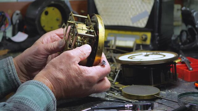 An elderly craftsman, in a workshop, repairs an old clock mechanism, an antique watch, adjusts gears, lubricates them with oil, cleans them from dust and dirt. Slow motion.