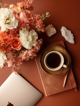 Elegant feminine workspace flat lay with flowers, coffee, laptop and journal on terracotta background