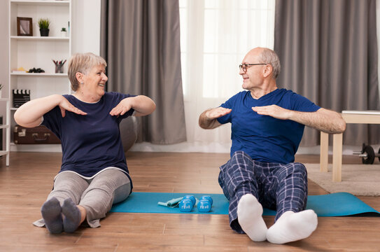 Aged couple stretches on a yoga mat and smiles at each other, sharing a home workout and healthy retirement routine, combining couple exercise, healthy aging and shared home workout routine.