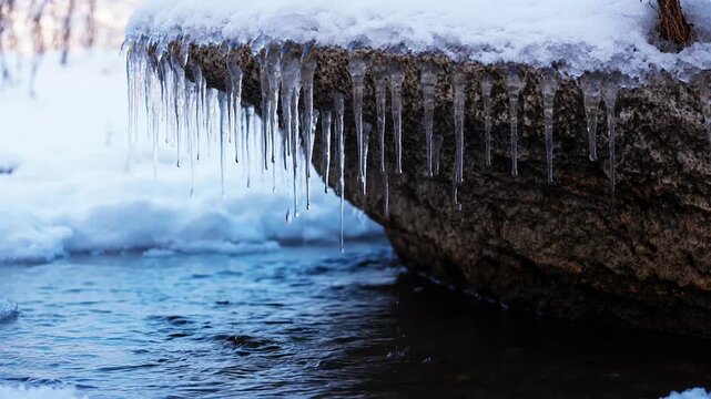 Clear icicles hanging from a rock formation above a cold flowing stream in a snowy winter landscape