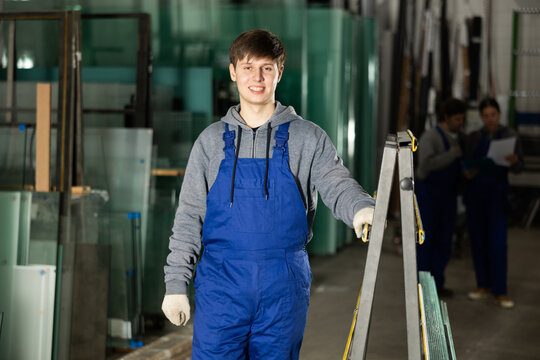 In glass workshop, young man glazer works with glass sheets and related supporting parts, preparing PVC products for interior finishing work.