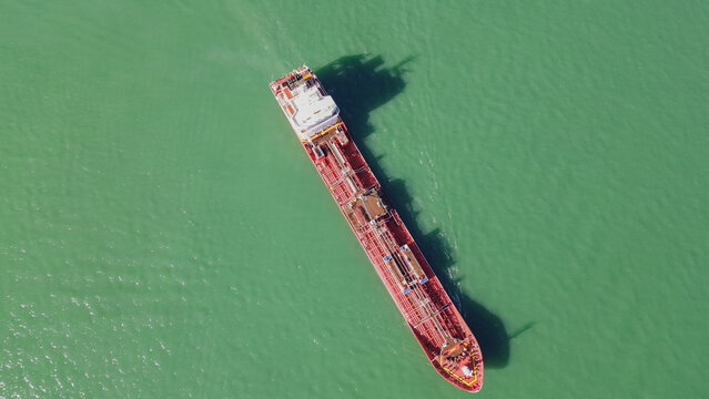 Horizontal aerial top down view of a massive red cargo ship navigating through the calm green ocean water with visible shadows on the surface of the sea