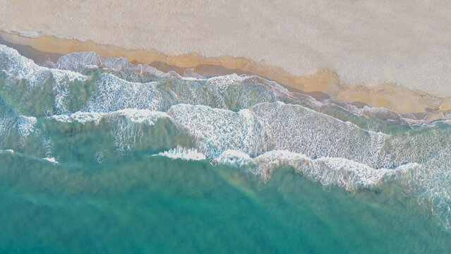 Horizontal aerial top down view of turquoise ocean waves washing on a sandy beach shore with sparkling white foam during a bright sunny summer day for travel promo