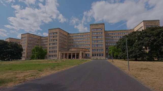 Grand symmetrical university building with beige stone facade, rows of windows, central portico, paved access road, grass lawn, mature trees, and bright cloudy sky
