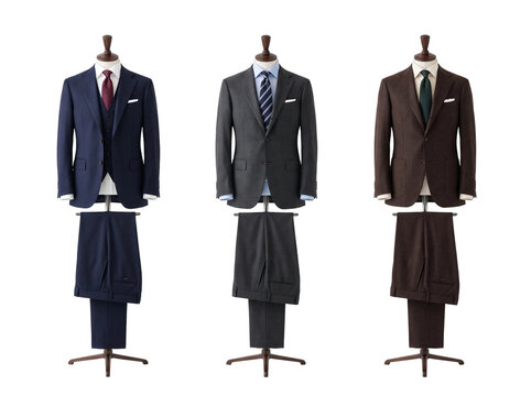 Three elegant professional mens suits featuring navy blue grey and brown formal tailored jackets with matching trousers white shirts and colorful neckties displayed on wooden store mannequins on trans
