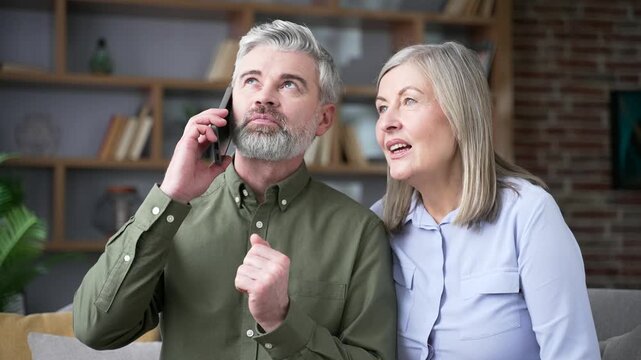 Close up of senior husband talking on a smartphone beside his wife while sitting on a sofa at home. Retired couple pleased with a recent service, sharing their experience and leaving a positive review