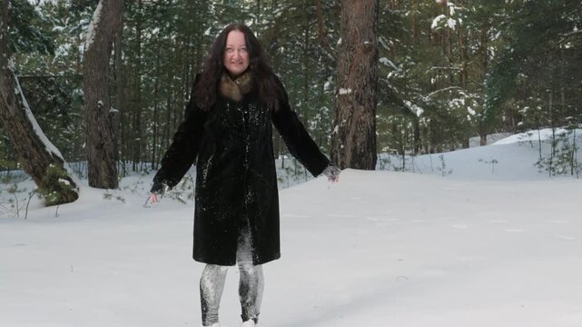 Woman in black coat trudges through deep snow surrounded by tall evergreen trees in winter landscape.