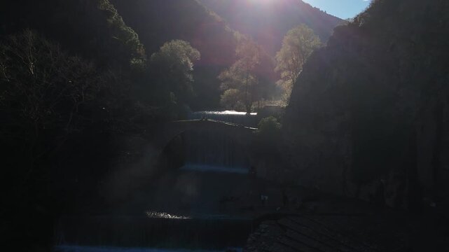 Cinematic drone pull back shot of a historic stone arch bridge and waterfall with dramatic sun rays
