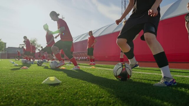 Womens soccer players practicing dribbling and ball control during organized training session on outdoor artificial turf field with cones under bright daylight