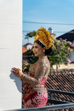 Beautiful Balinese bride in traditional wedding attire with golden crown payas agung and elegant lace kebaya, smiling on a balcony under bright blue sky.