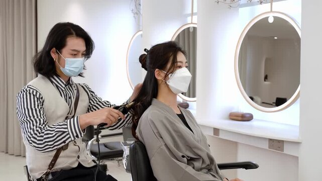 Hairdresser stylist making curls a luxurious evening hairstyle with a curling iron and make wave for long hair of the female customer with silk robes in modern spa hair salon care service for stylish