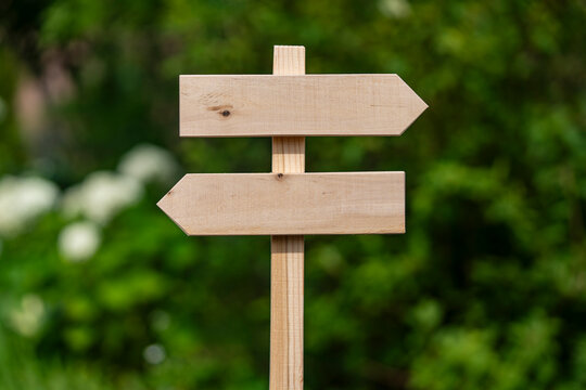 Empty wooden sign post with two arrows pointing in opposite directions on blurred green garden background for copy space business decisions navigation and marketing choices