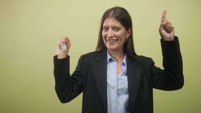 Woman holding a house key fob and pointing finger in studio with green backdrop and blazer; confidence success.