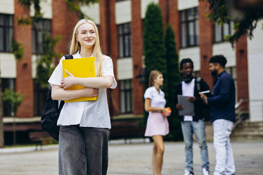 Young blonde student embracing education, holding a yellow folder and a backpack while standing on a diverse university campus