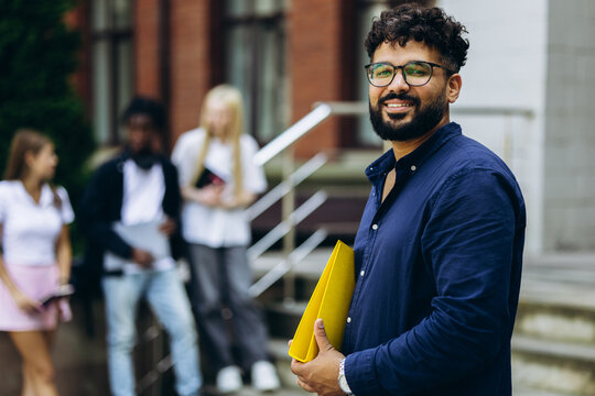 Smiling male student holding a yellow folder on university campus with diverse students in background, representing education and youth